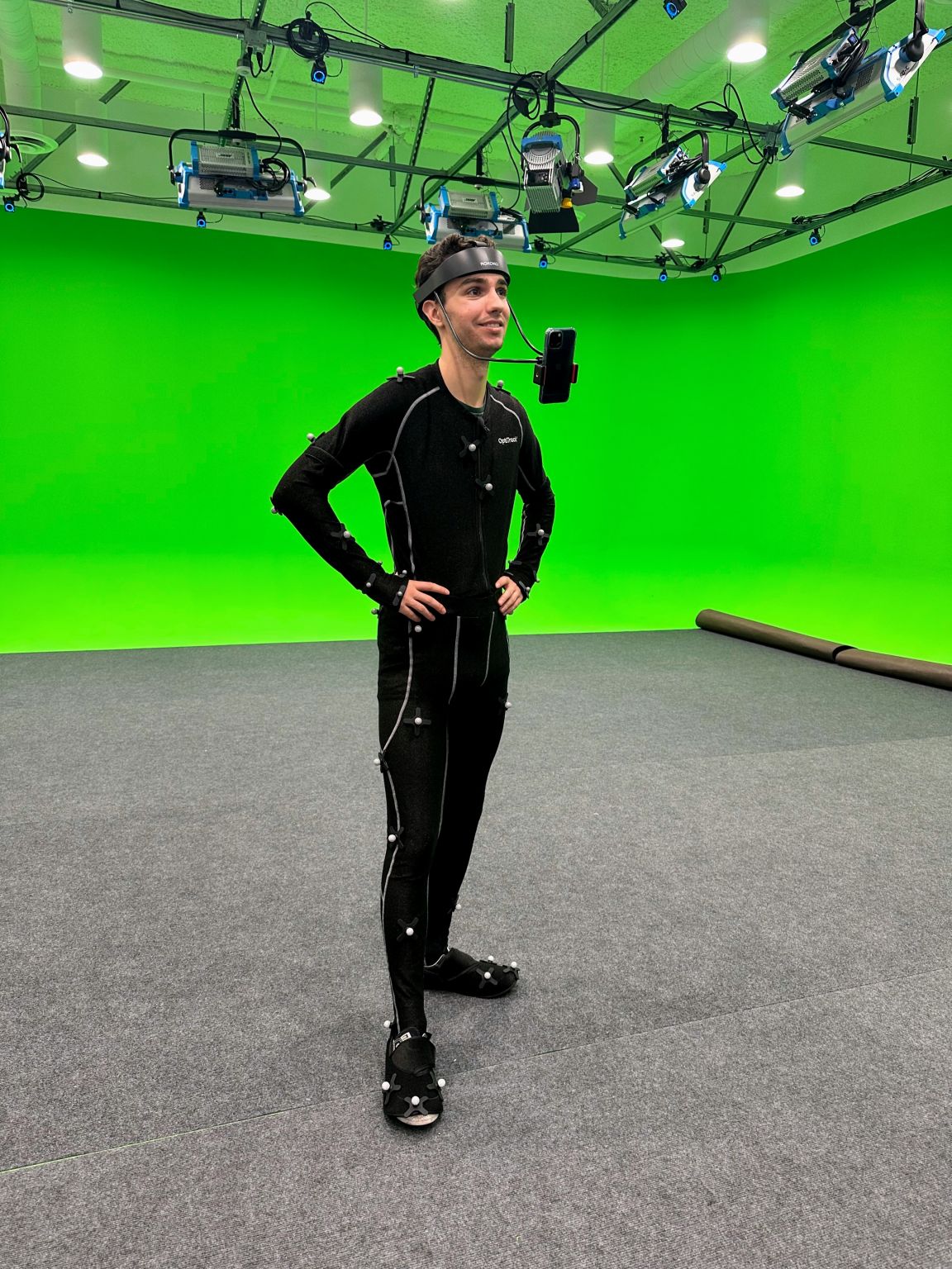 Andrew in an OptiTrack motion capture suit with reflective markers and a head-mounted phone rig, standing on a green-screen stage under studio lights